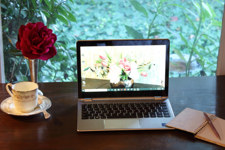 Silver laptop on dark wood desk in front of a window. Green bushes outside, a red rose in a silver vase, and a teacup on a saucer to the left. Small notebook an pen at right.