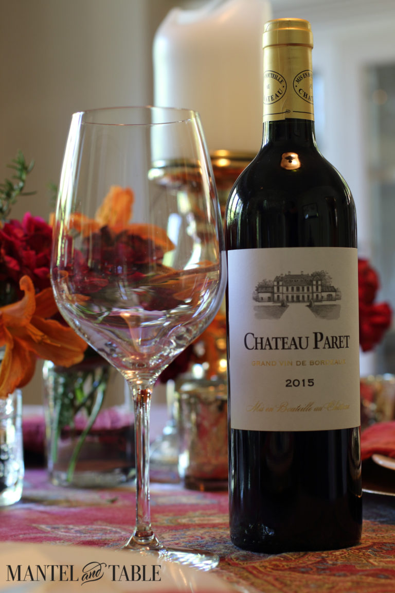 Chateau Paret wine bottle with glass in front of orange & red flowers & white candle