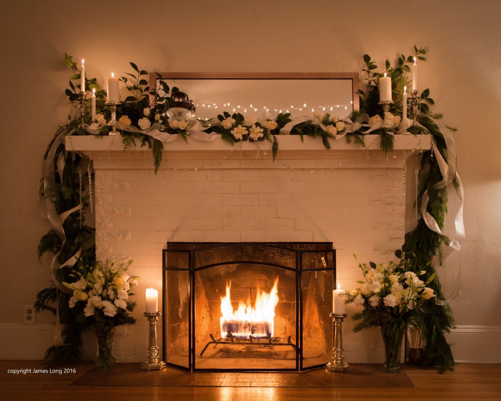 Fire in the fireplace. Evergreen garland with white roses, gardenias, & lilies, candles, on the mantle, and vases of white flowers on the hearth.