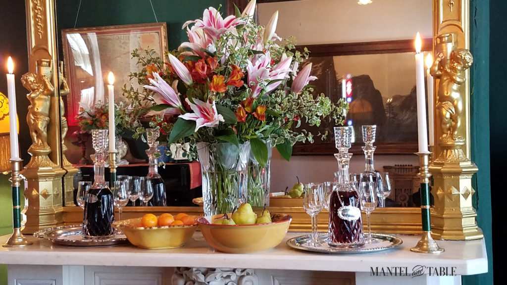 Beautiful white marble mantel with large flower arrangement of lilies, with bowls of fruit, and cut glass port and sherry decanters on trays. Gold gilt mirror in the back.