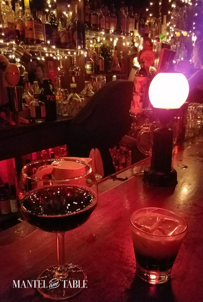 The reddest bar ever - a glass of red wine & a shot of something on the bar, with a small lamp & lots of twinkle lights in the background.