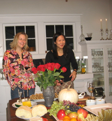 Two lovely ladies holding red wine glasses, at the food table with red roses, a white pumpkin, fruit tray, cheese plate, & other yummy things to eat!