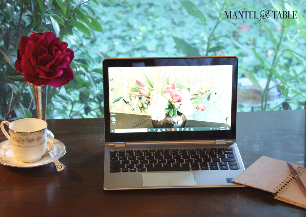 Silver laptop on dark wood desk in front of a window. Green bushes outside, a red rose in a silver vase, and a teacup on a saucer to the left. Small notebook an pen at right.