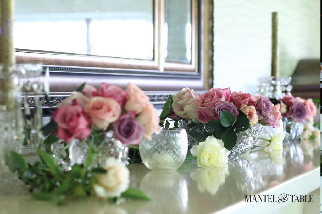 Angle view of the mantel with pink roses in crystal vases, cream & soft yellow roses lying on the mantel, & votive candle holders.