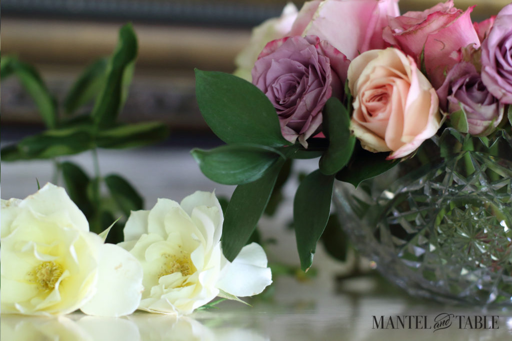 Two soft yellow garden roses lying on the mantel to the left of a crystal bowl of pastel roses. A green rose bough lies behind.