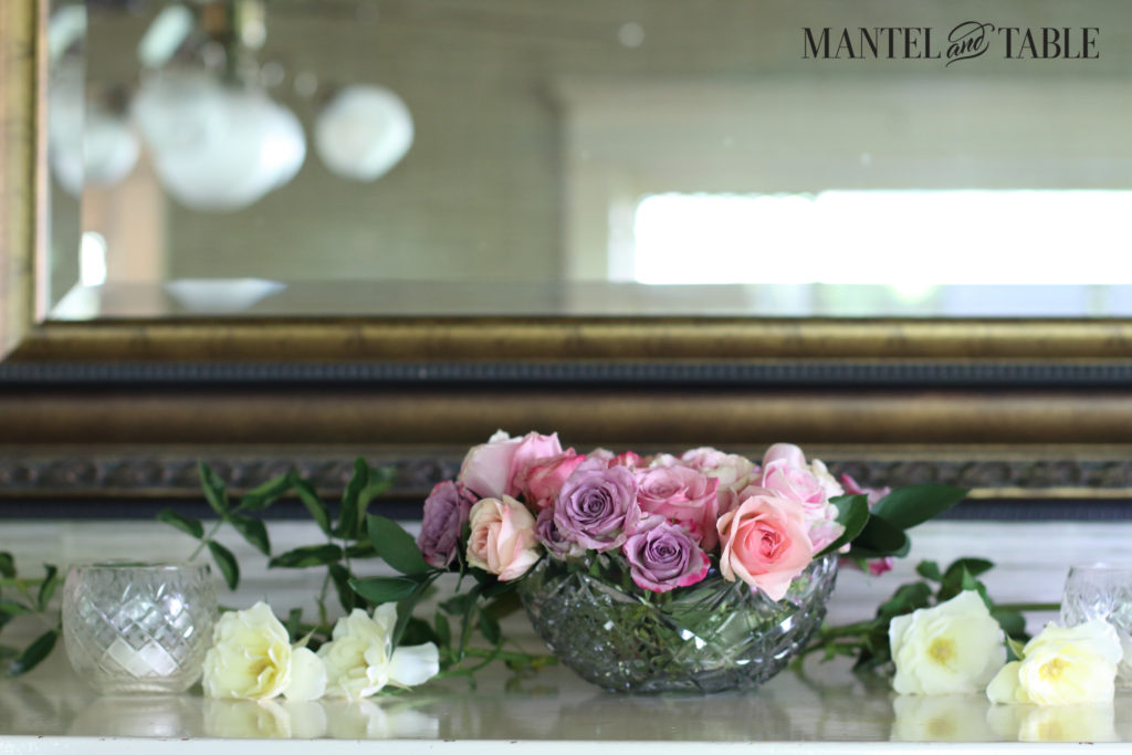 Pastel roses in a crystal bowl flanked by soft yellow garden roses lying on the mantel. Lovely wood & gold mirror in the background.