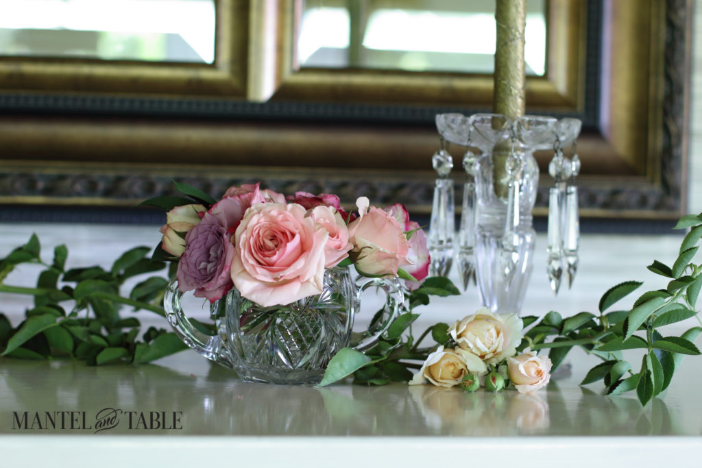 Green rose bough twining through the pastel rose arrangements, crystal candle holder at the right.