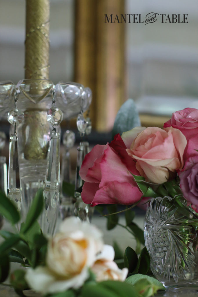 Crystal candle holder with gold candle, slightly deeper pink roses in front, and cream roses lying on the mantel.