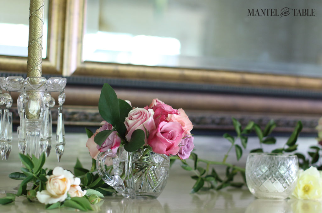 A long rose leaf bough lies on the mantel in back of cream roses, & pink roses in crystal vase. Gold candles in a crystal holder to the left.