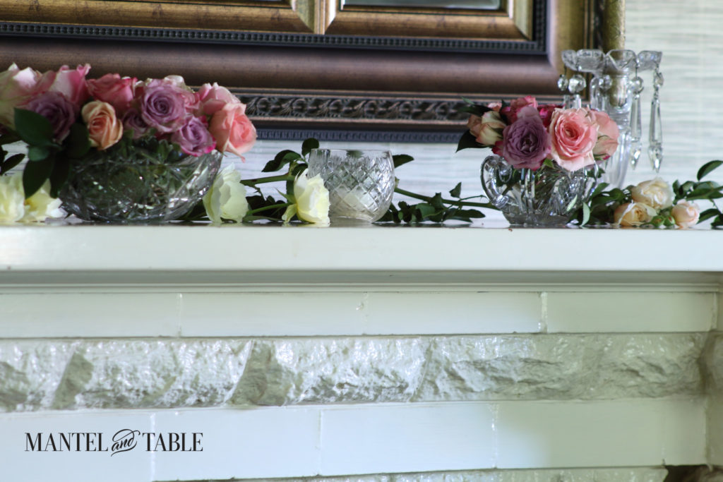 Pastel pink & lavender roses in crystal bowls bookending soft yellow garden roses lying on the mantel, crystal chandelier candle holder on the right.
