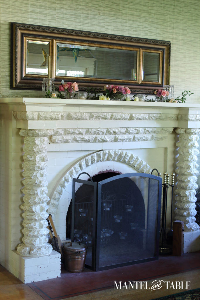 Full view of the fireplace, white painted brick with textured rows, pillars at the sides of the arched opening, and a lovely antique mirror horizontally across the top.