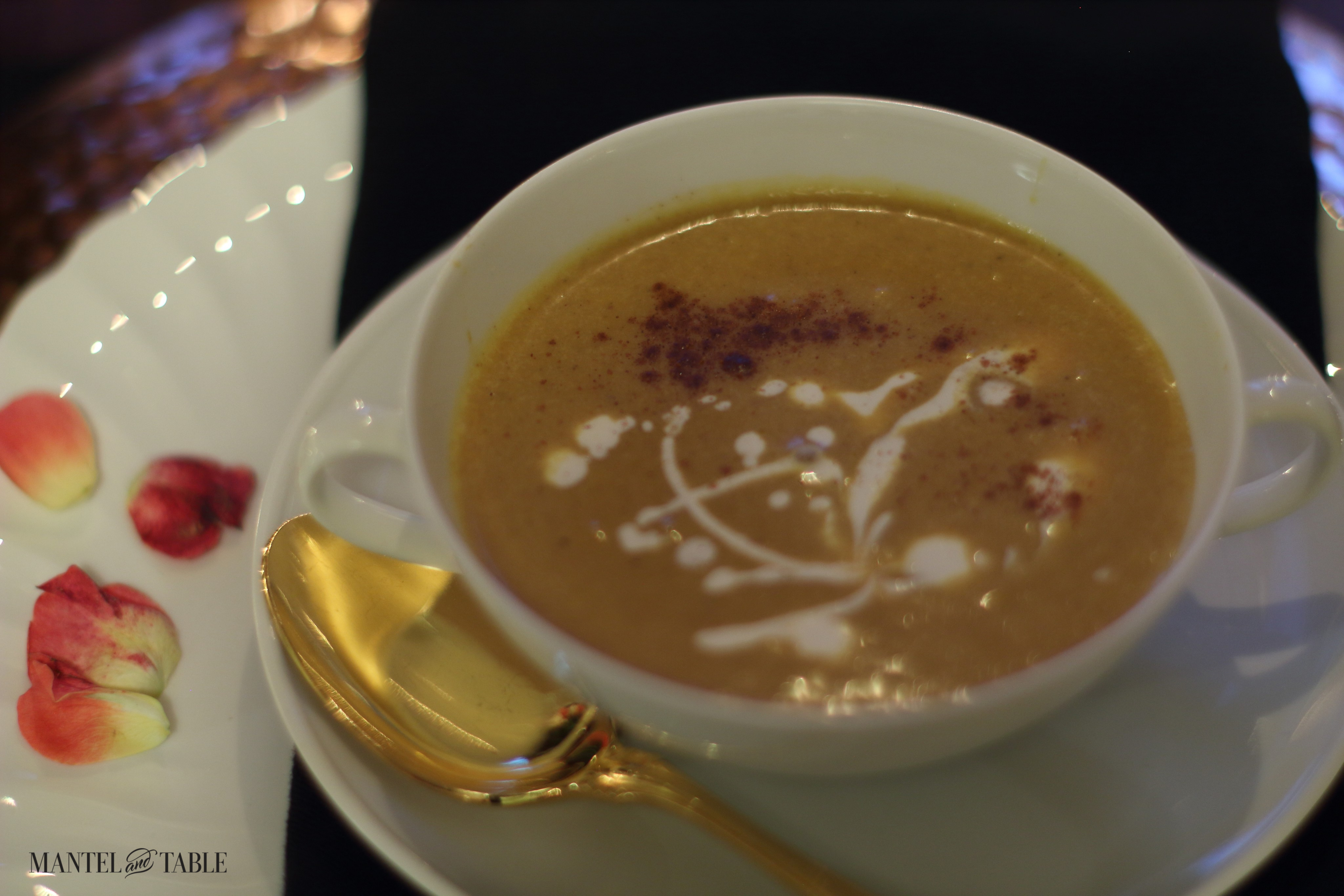 Pumpkin soup in a white two-handled bowl, garnished with heavy cream and cinnamon. Gold spoon on the saucer, foreground