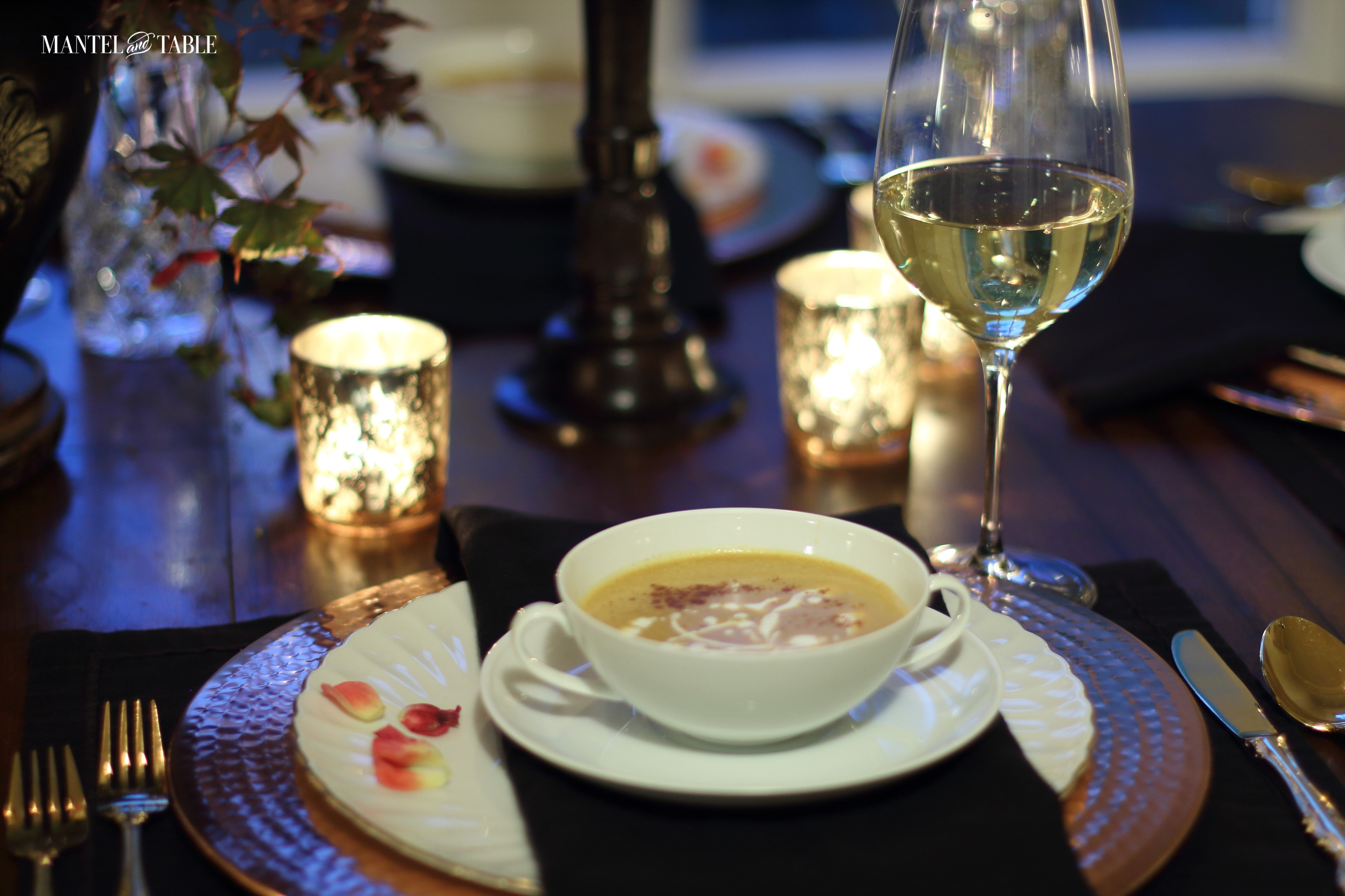 Place setting with white plates, black napkin, copper charger, and white soup bowl full of pumpkin soup! Glass of white wine at the right.