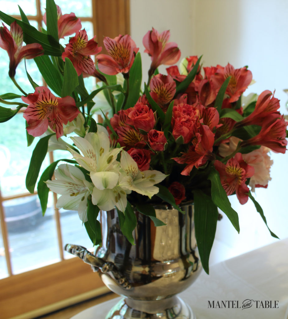 Light pink carnations, deep pink baby roses, pink & white altromeria in two-handled, silver wine bucket on a white tablecloth