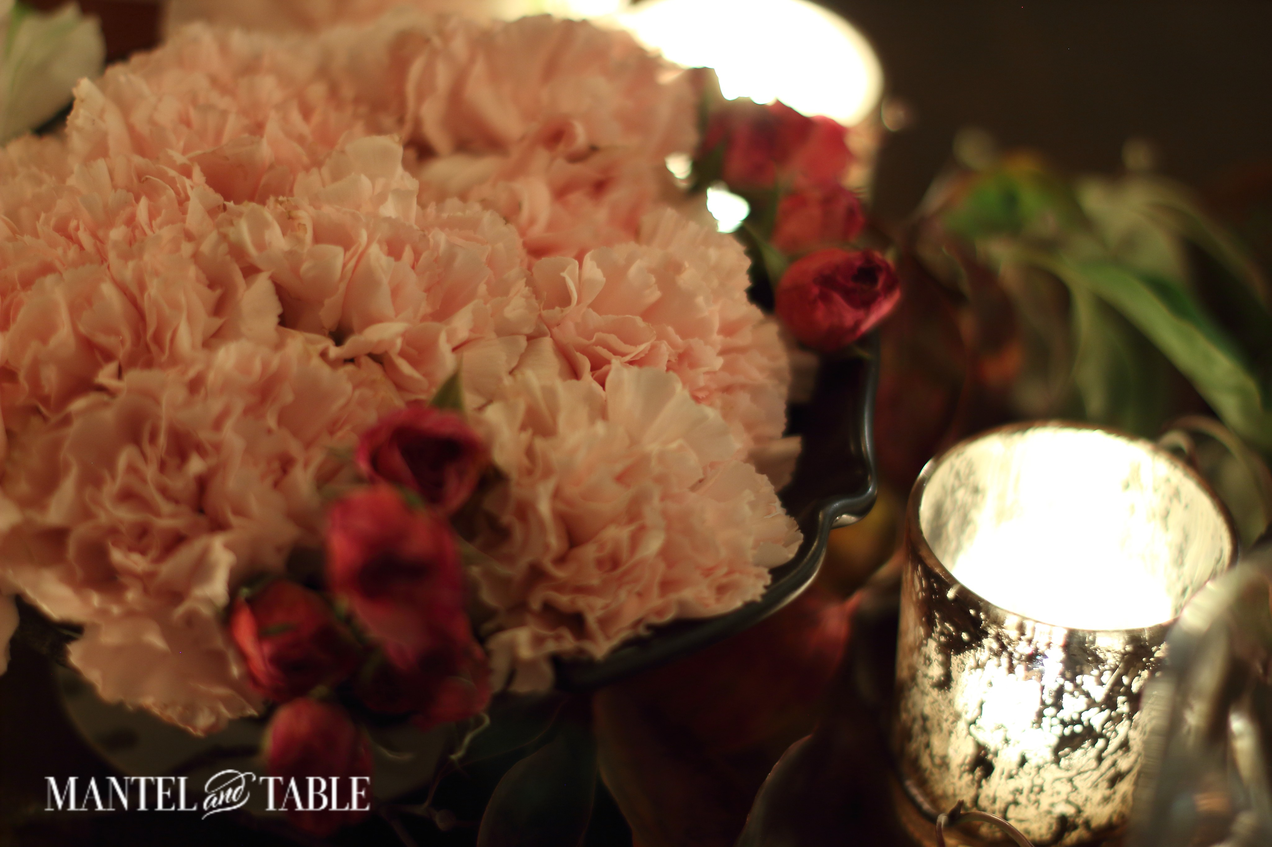An arrangement of pInk carnations and deep pink baby roses beside a glowing brass-toned votive and fall leaves. on a copper table