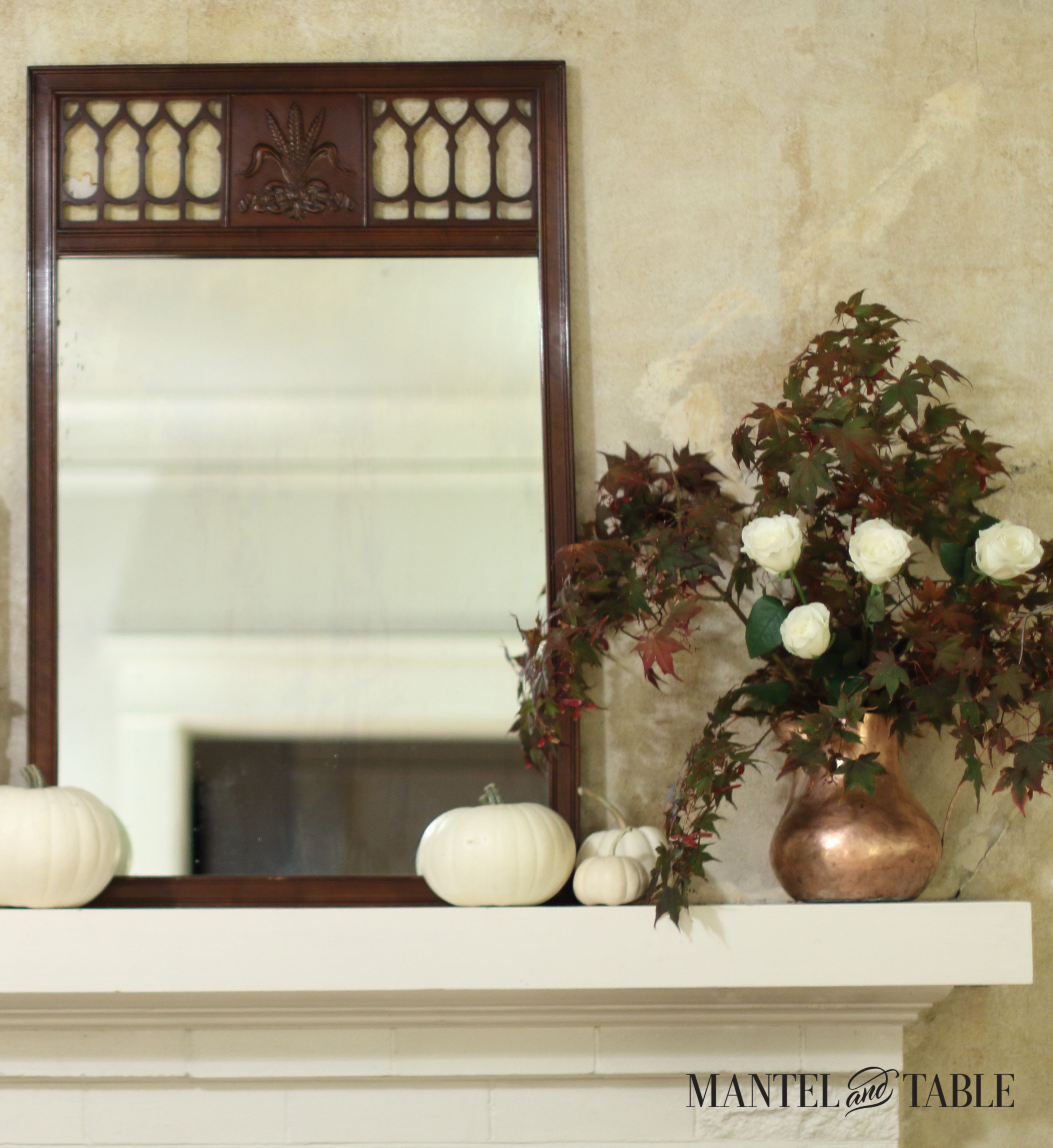closeup of tall dark wood mirror on white mantel. White pumpkins in front, and copper pitcher full of fall leaves & white roses on the right.