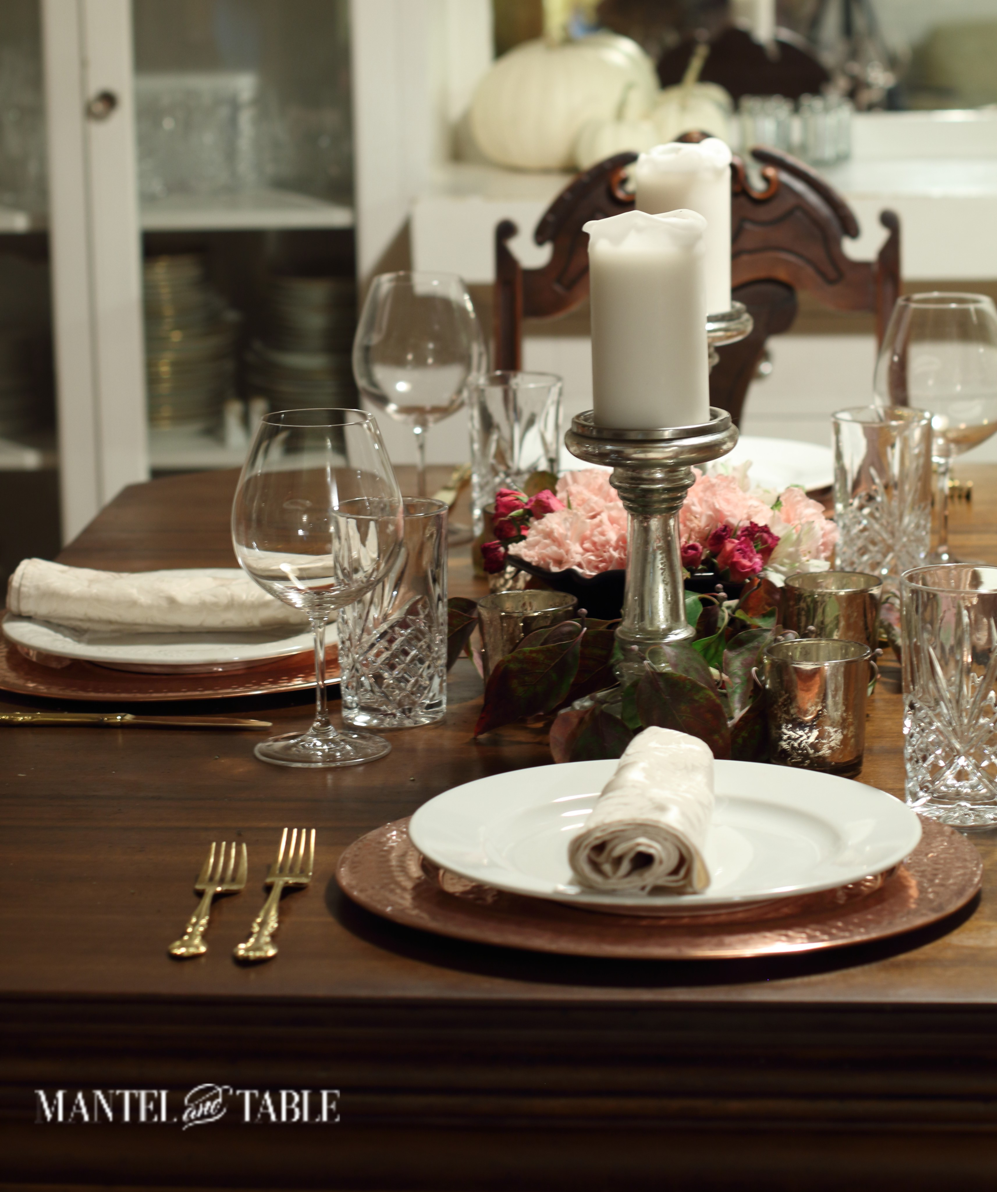 Dark wood table with copper chargers, white plates, silver candle holders, pink flower & fall leaves centerpiece.