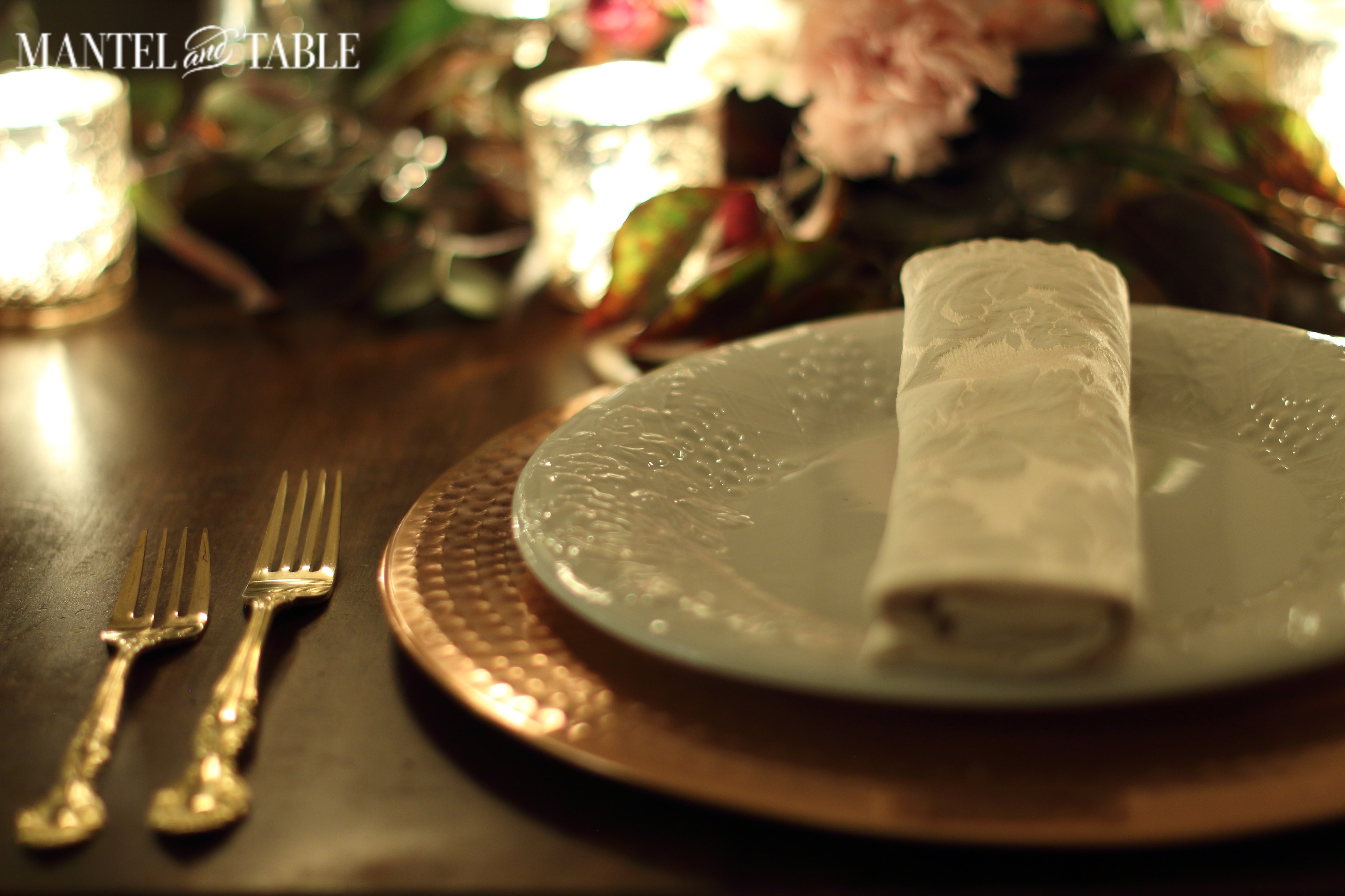 Autumn colored leaves behind the copper charger, white plate & blush napkin stack. Gold flatware to the left, and glowing votive candles tucked in the leaves.