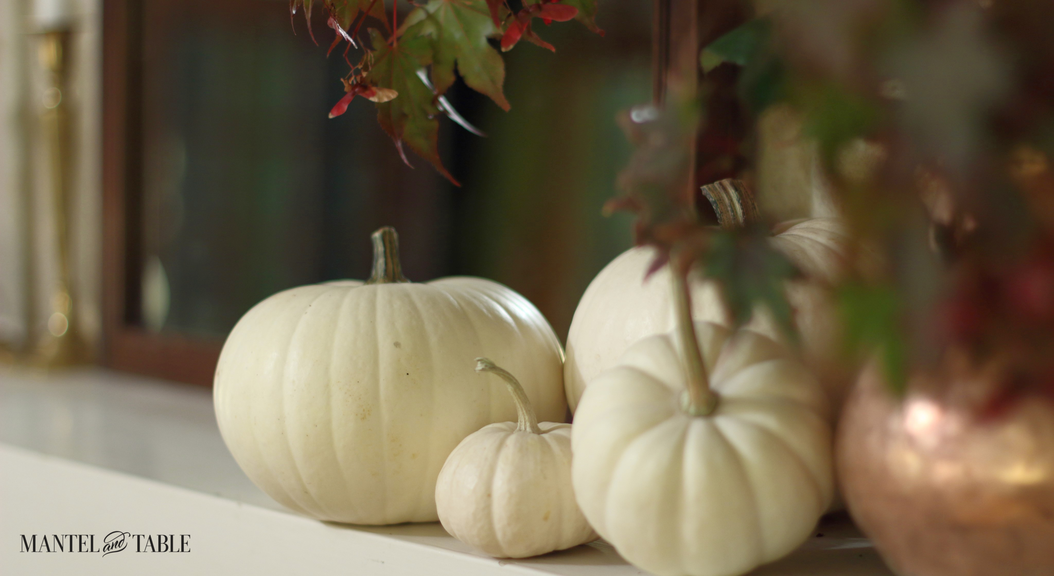 Closeup Group of white pumpkins on white mantel next to copper pitcher with fall leaves.