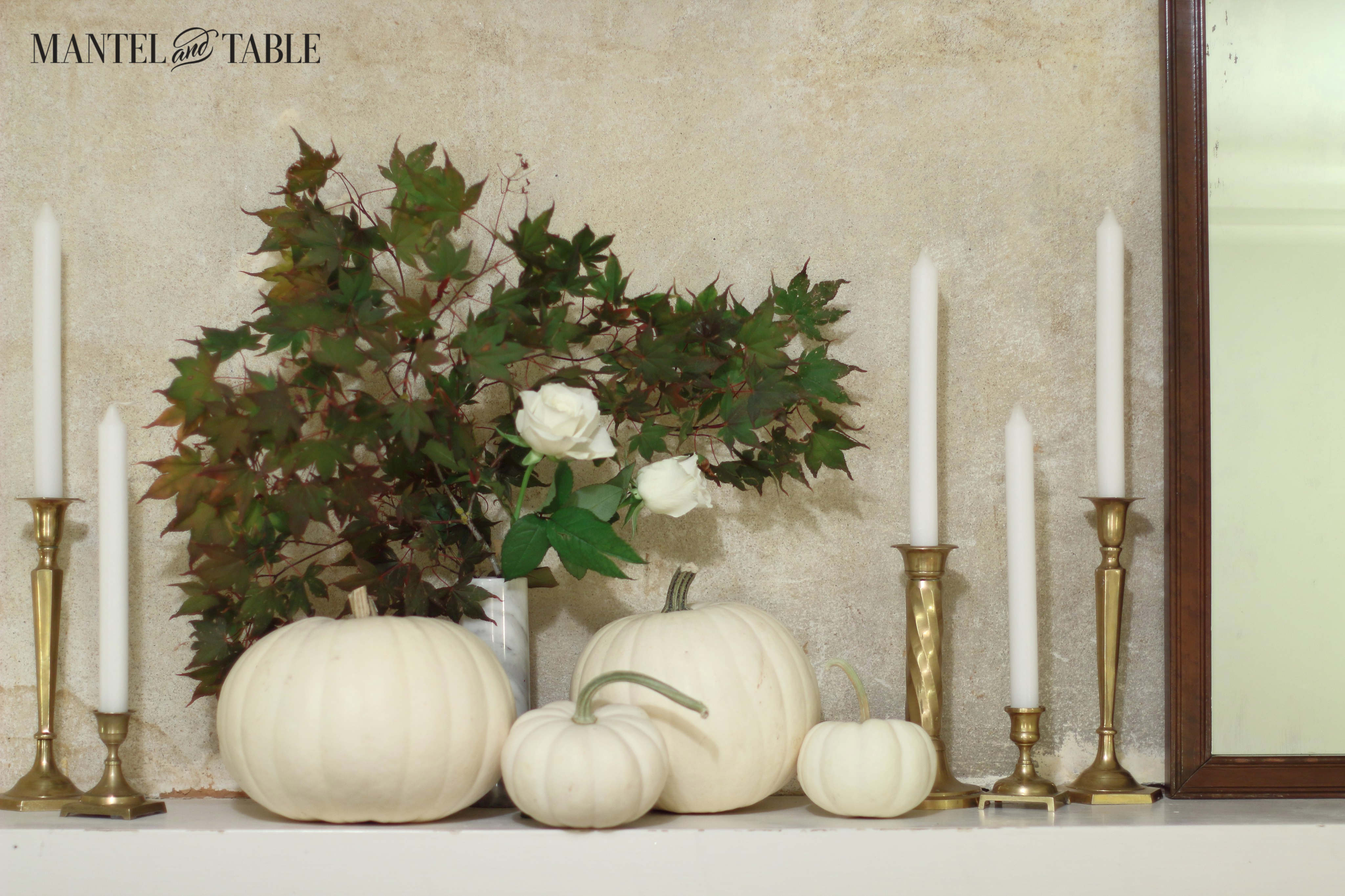 White pumpkins on white mantel, with white marble vase behind, full of fall leaves &: white roses. Brass candle sticks & white candles on the right.