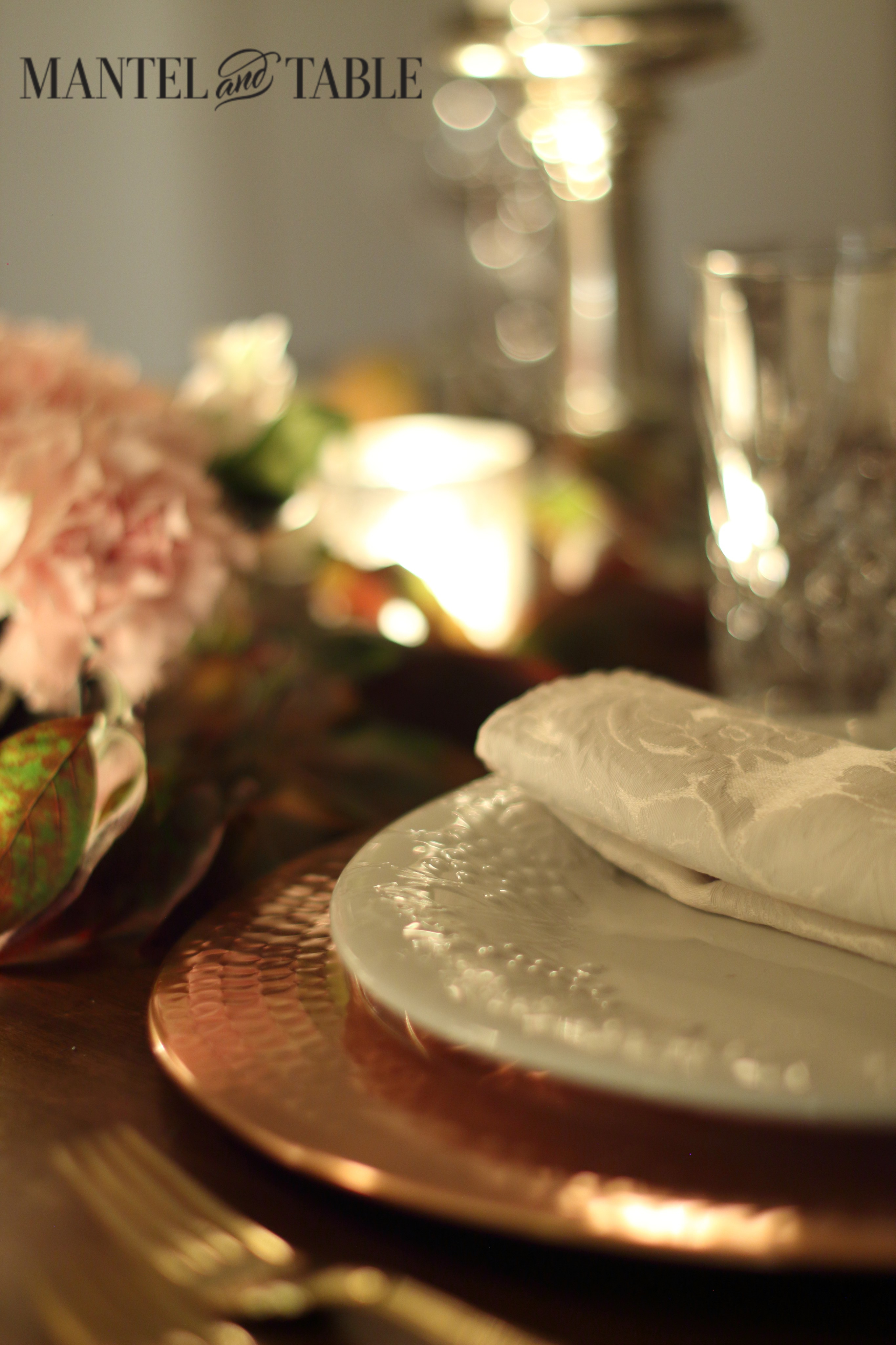 Copper charger, white figured plate, blush napkin with cut glass and silver candle holder at the back right, pink centerpiece to the left and glowing brass mercury glass votive in the middle.