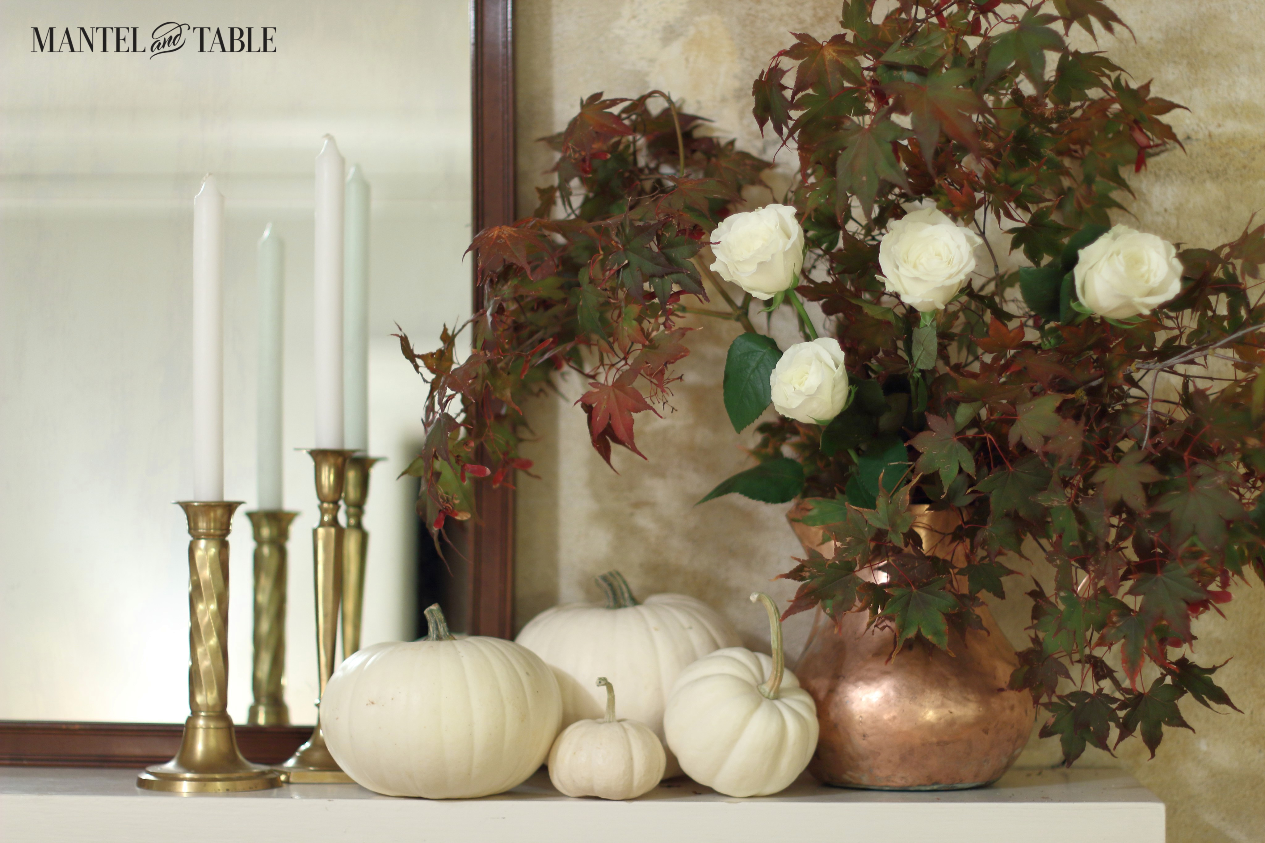 White pumpkins on the mantel. Copper pitcher with fall leaves & white roses on the right, brass candle sticks & white candles on the left in front of a dark wood mirror.