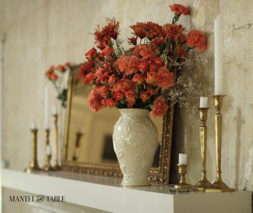 White mantel at an angle, cream vase of peach & rust flowers, brass candlesticks & gold framed mirror