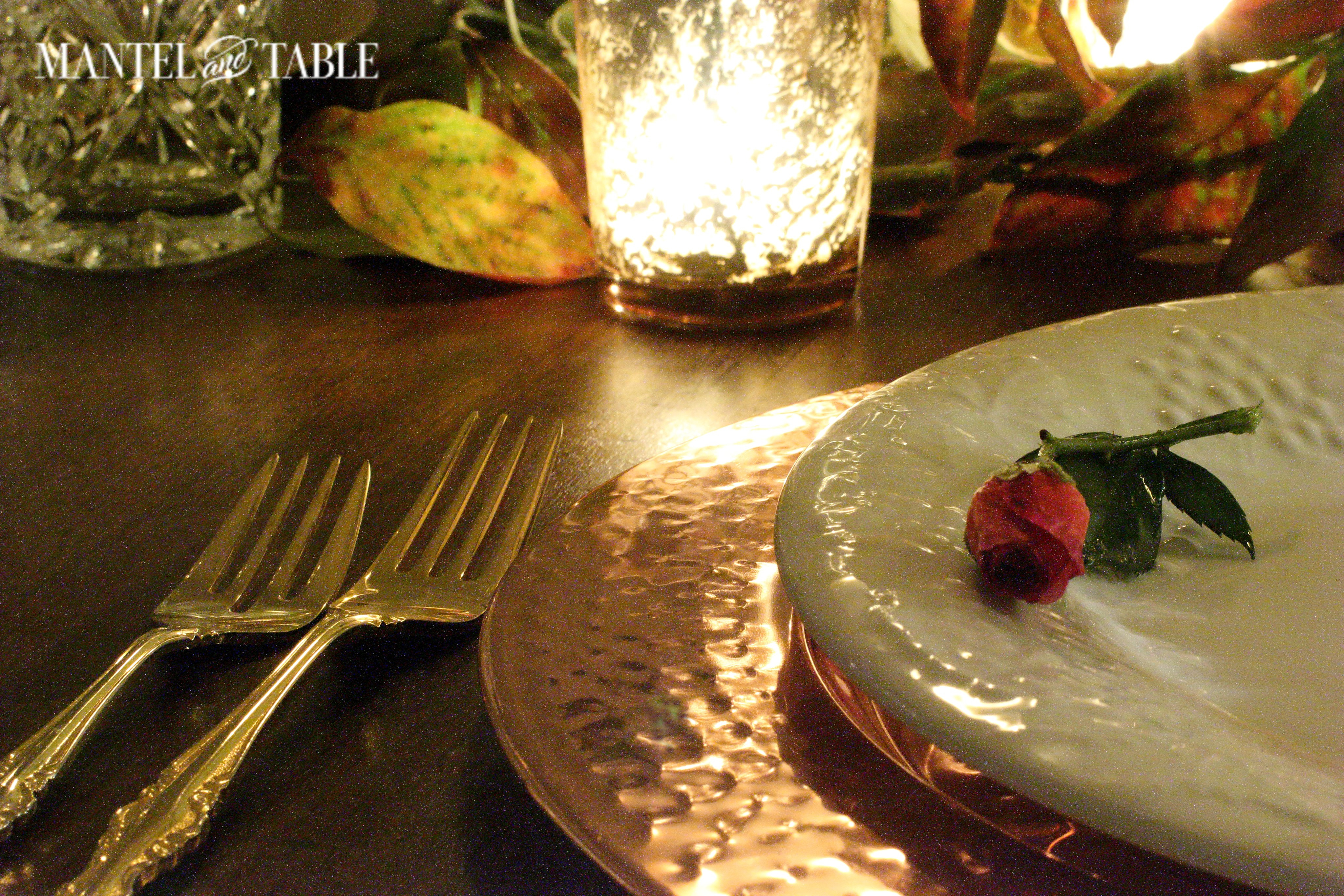 Closeup of hammered copper charger with white plate on top. A deep pink rose on the plate. Gold forks to the left and a brass mercury glass votive above.
