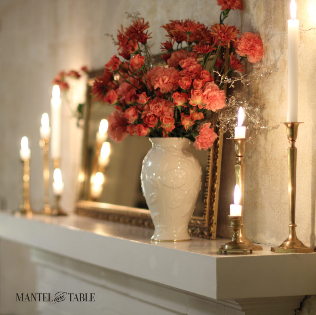 White mantel at an angle, cream vase of peach & rust flowers, glowing brass candlesticks & gold framed mirror