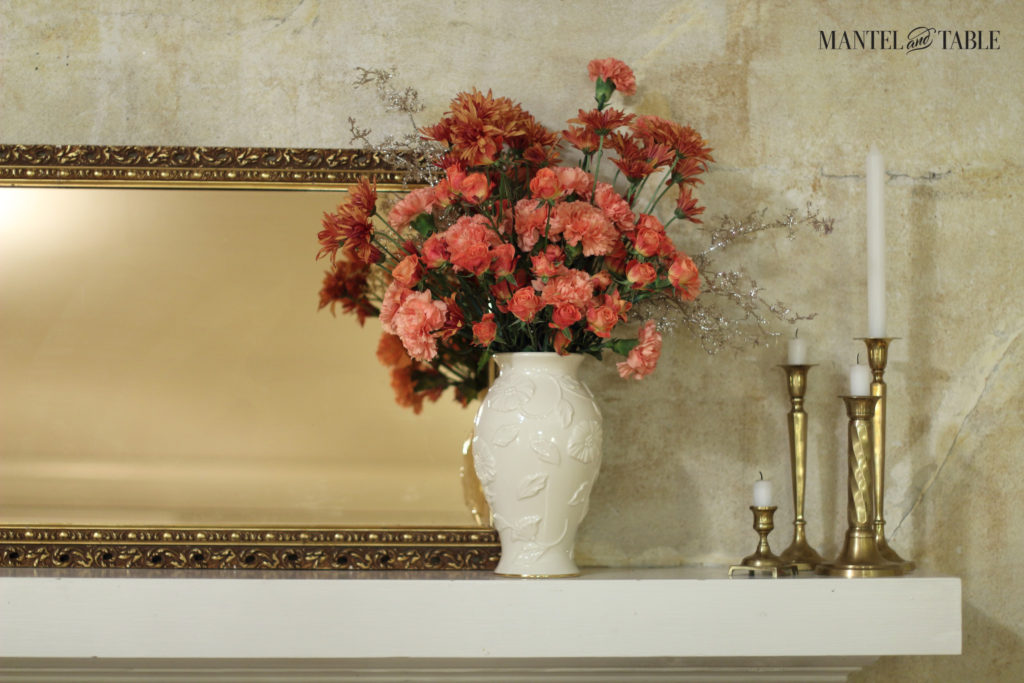 White mantel. 4 brass candlesticks of various heights to the right. Peach flowers in white vase, and gold framed mirror behind.