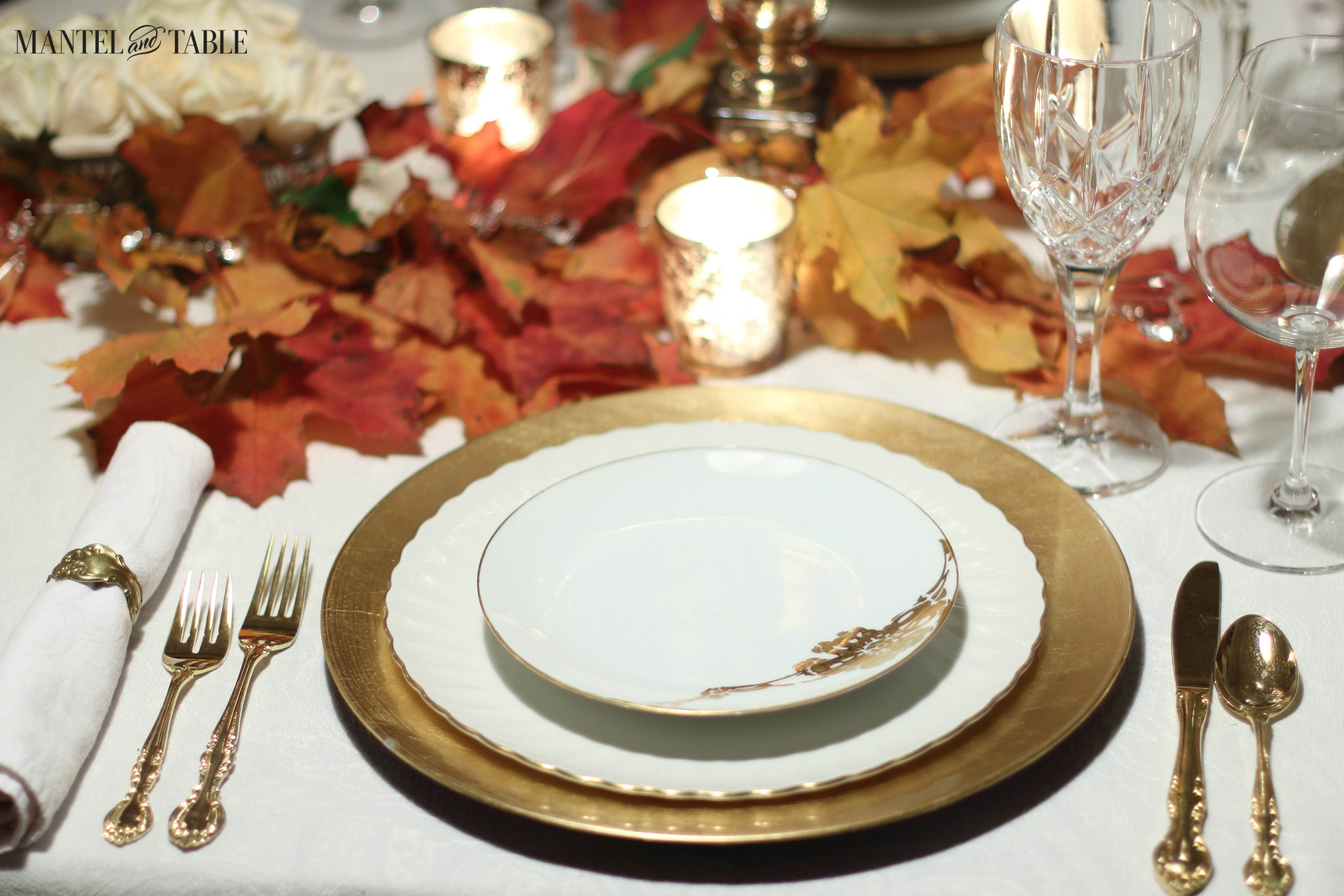Gold & white plate stack with gold tableware. Red & orange leaves above the plates, with a glowing gold mercury glass votive.