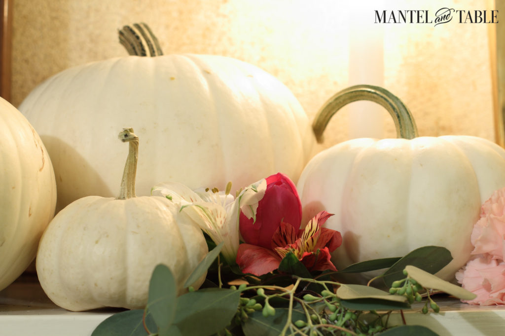 White pumpkins with seeded eucalyptus & pink flowers in front. Candle glow in back.