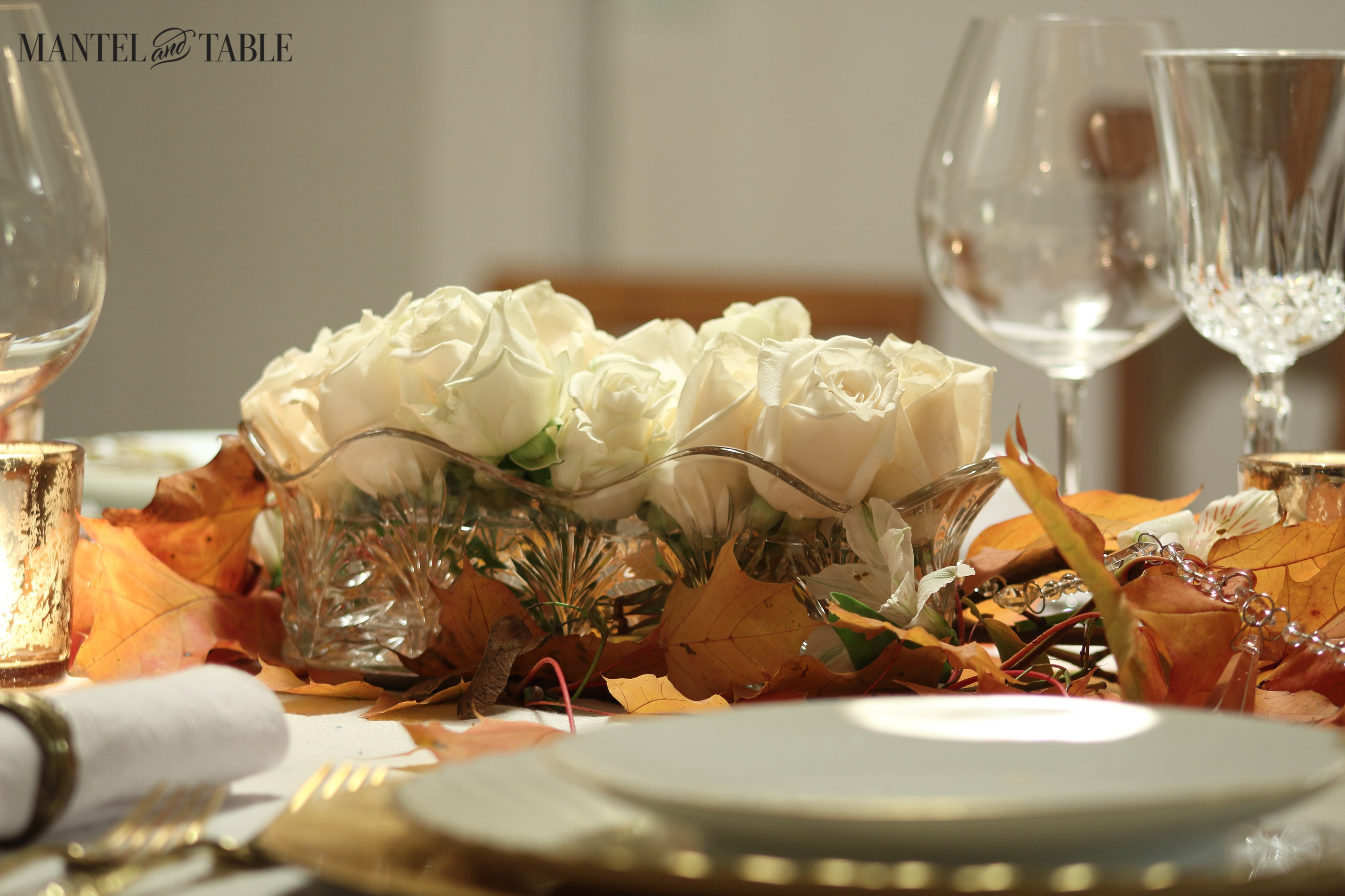 White roses in a long cut glass vase surrounded by fall leaves