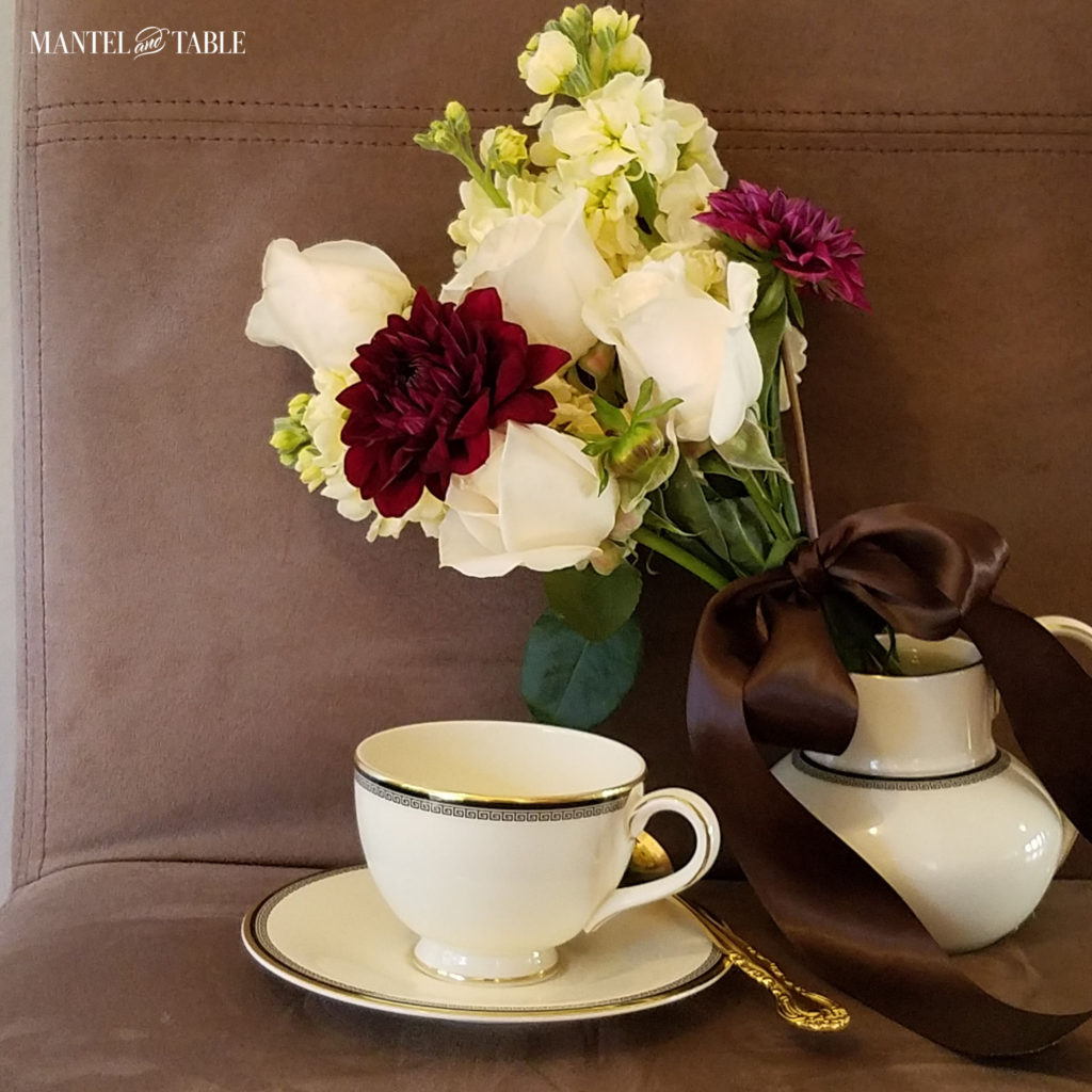cream & gold greek key trim teacup on brown suede chair with white rose & burgundy mum bouquet tied with brown satin ribbon