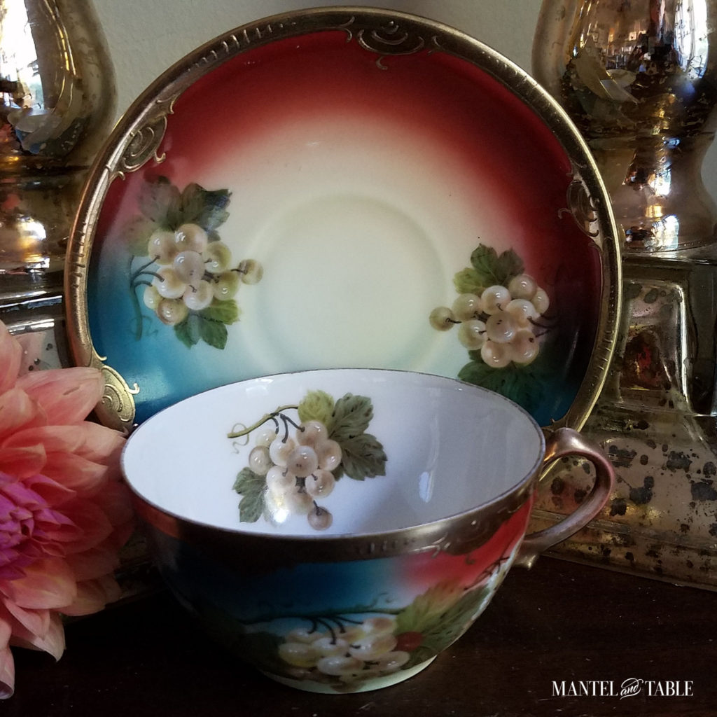 bright turquoise & rust teacup with gold trim. & handpainted white grapes Between two gold pillar candle holders, peach mums