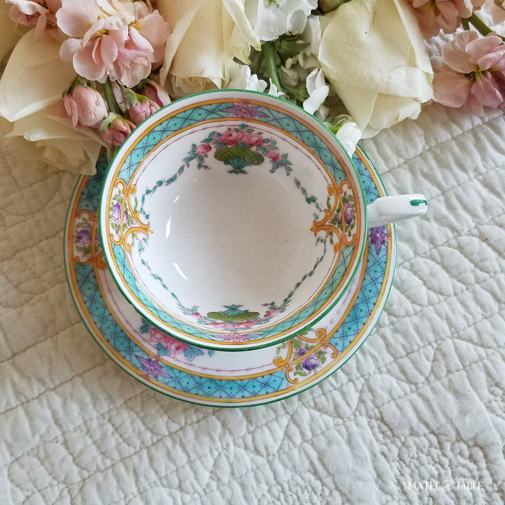 white teacup with teal green trim & pink & gold detail on white quilt, pale pastel flowers