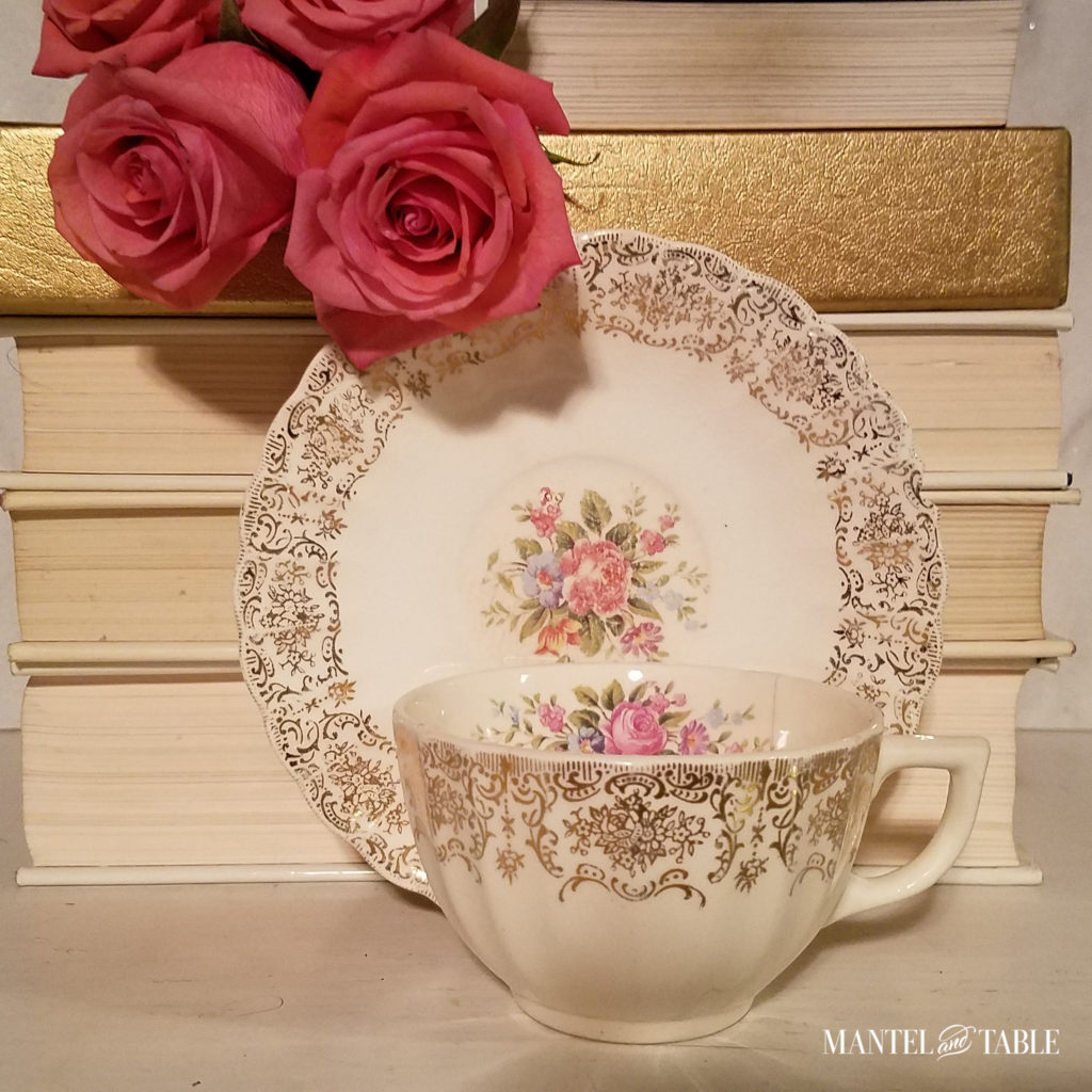 cream teacup with gold filigree trim, light pink & blue flower pattern in the center, leaning against stack of white books, with salmon pink roses