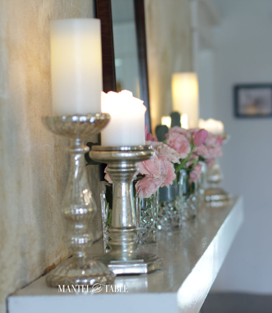 Silver pillar candleholders foreground, pink flowers in crystal vases on mantel behind