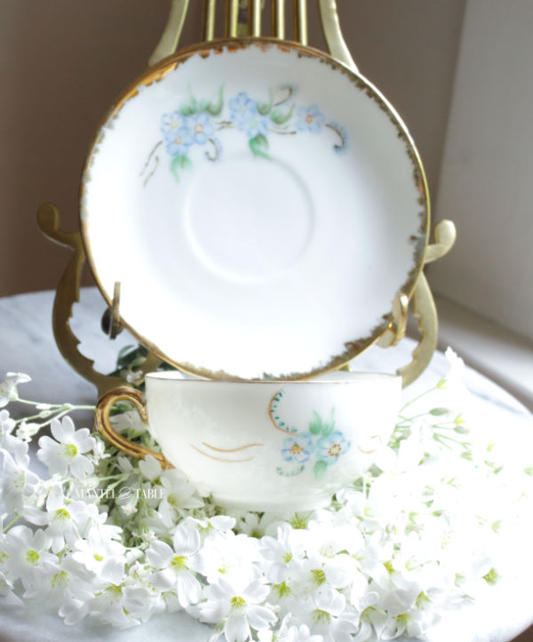 White saucer on brass stand with Snow on the Mountain fresh flowers under the teacup.
