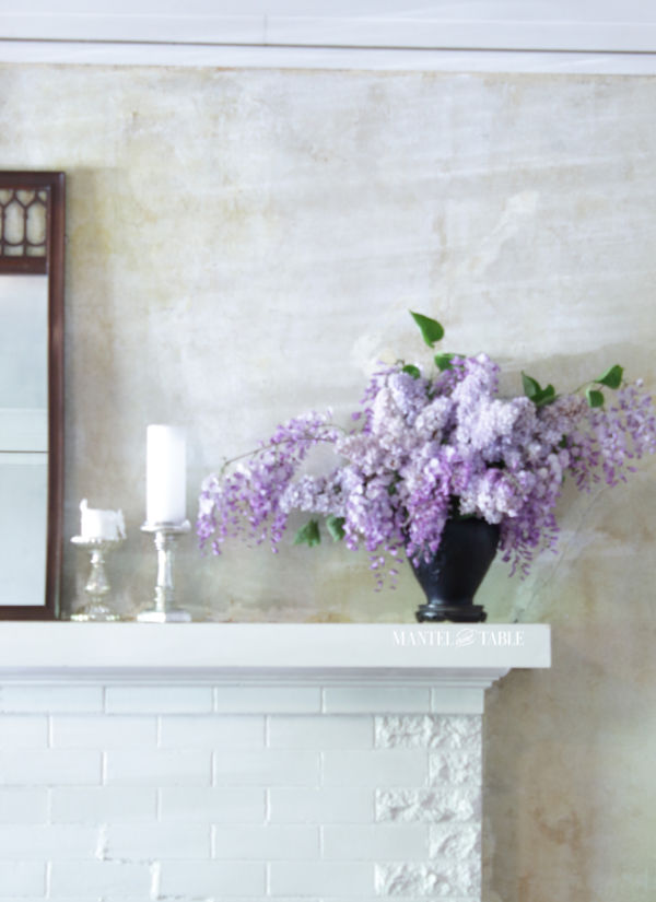 Black amethyst glass vase with lavender lilacs and wisteria on the corner of a white brick mantel with silver candle holders and a brown wood mirror