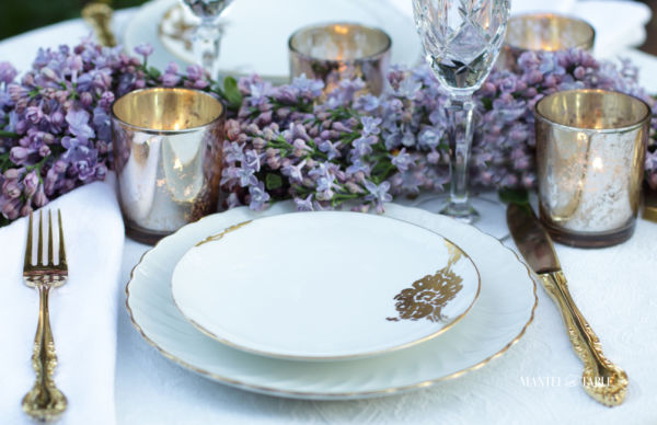 White and gold china place setting with gold votives, gold flatware and lilacs.