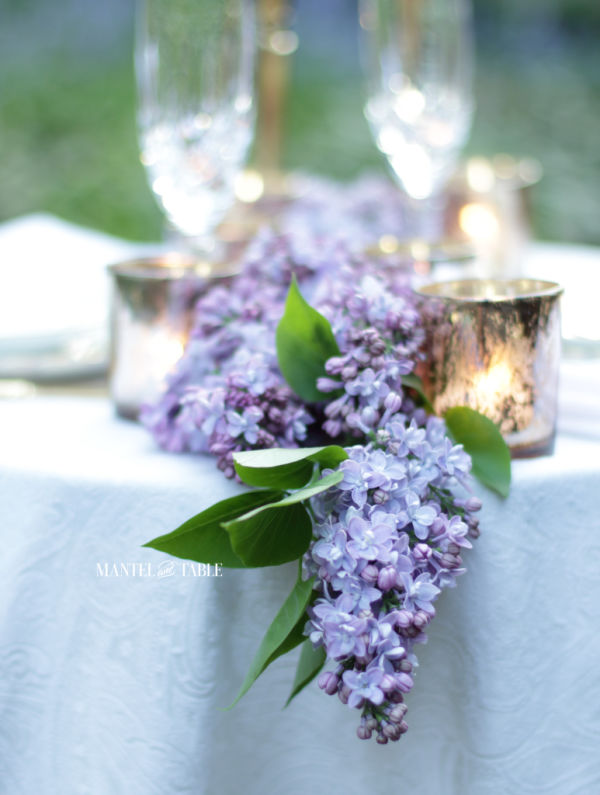 Beautiful light purple lilac garland overhanging white tablecloth on outdoor dinner table for two.