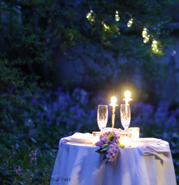 White tablecloth and candles on outdoor dinner table for two with lights in the trees above.