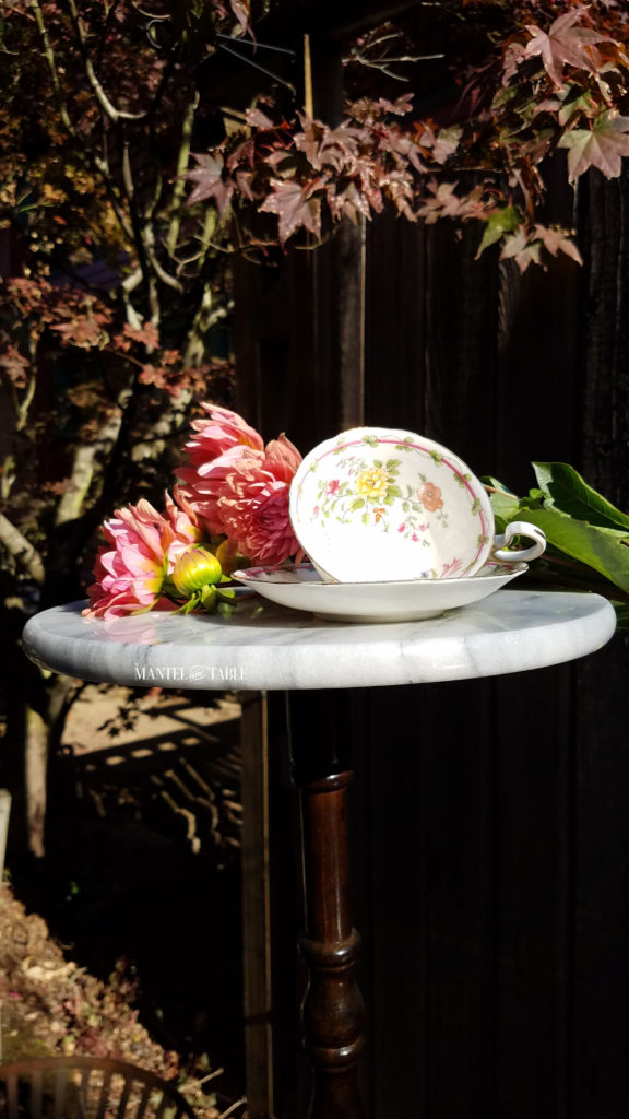Teacup and saucer. White with fall colored flowers on gold stand. Sitting on white marble base with peach dahlias against a rough wood fence background.