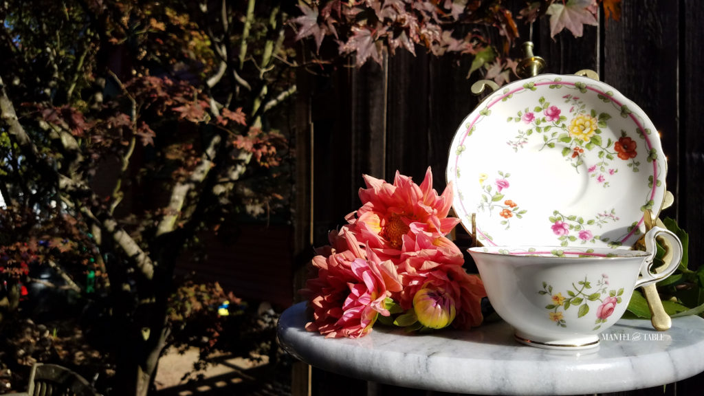 Teacup and saucer. White with fall colored flowers on gold stand. Sitting on white marble base with peach dahlias against a rough wood fence background.