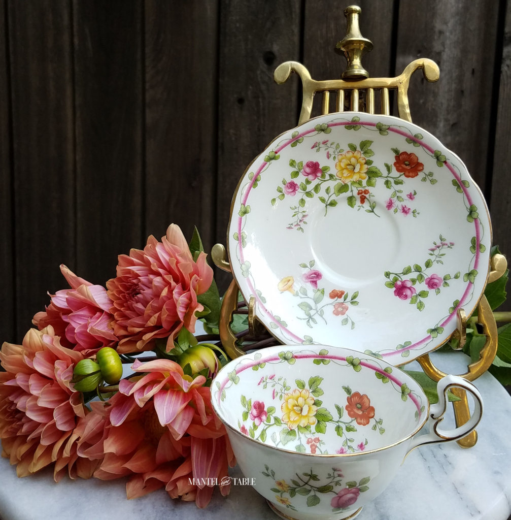Aynsley China teacup and saucer. White with fall colored flowers on gold stand. Sitting on white marble base with peach dahlias against a rough wood fence background.