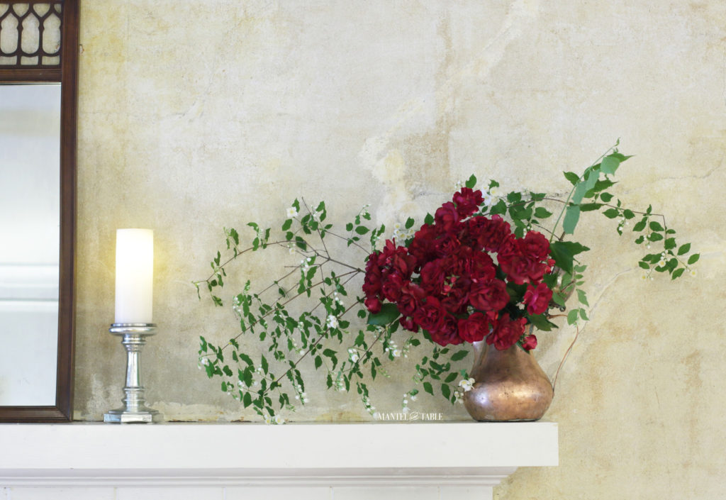 Red roses, mock orange branches in a copper pot on white mantel. Silver candle holder and edge of brown filigree mirror.