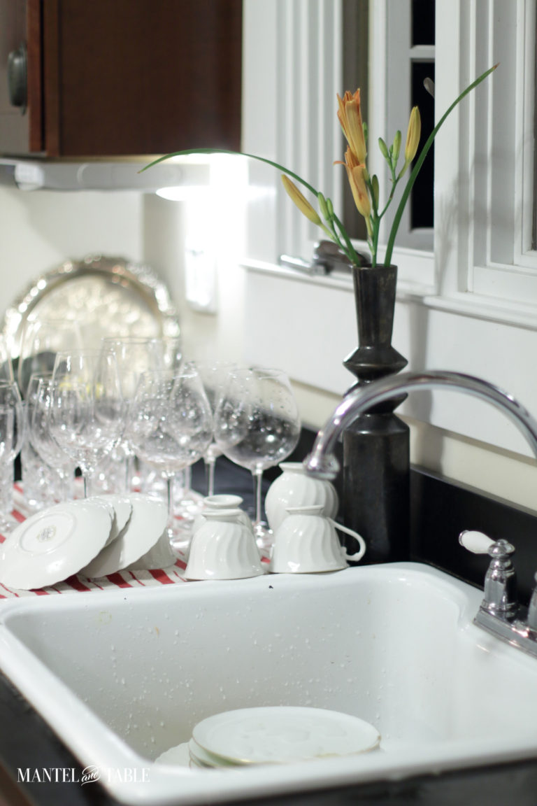 Glasses and china drying beside the sink. Orange daylily in a vase and string lights outside the window during clean up