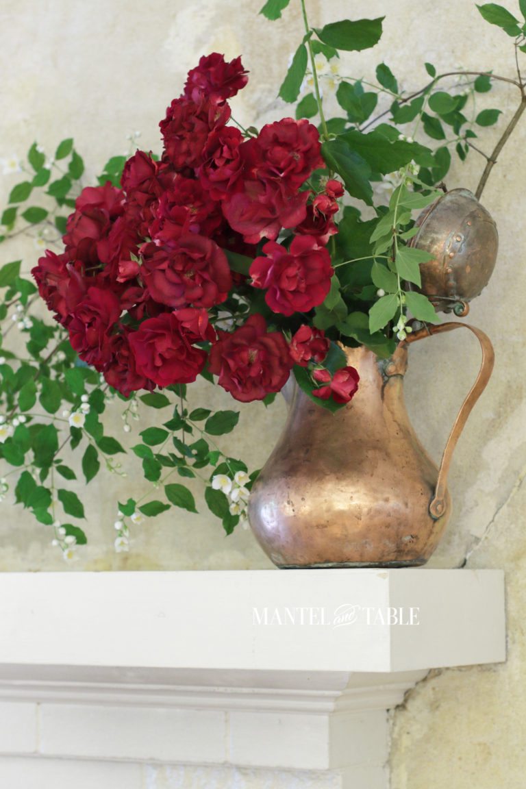 Red roses, mock orange branches in a copper pot on white mantel.