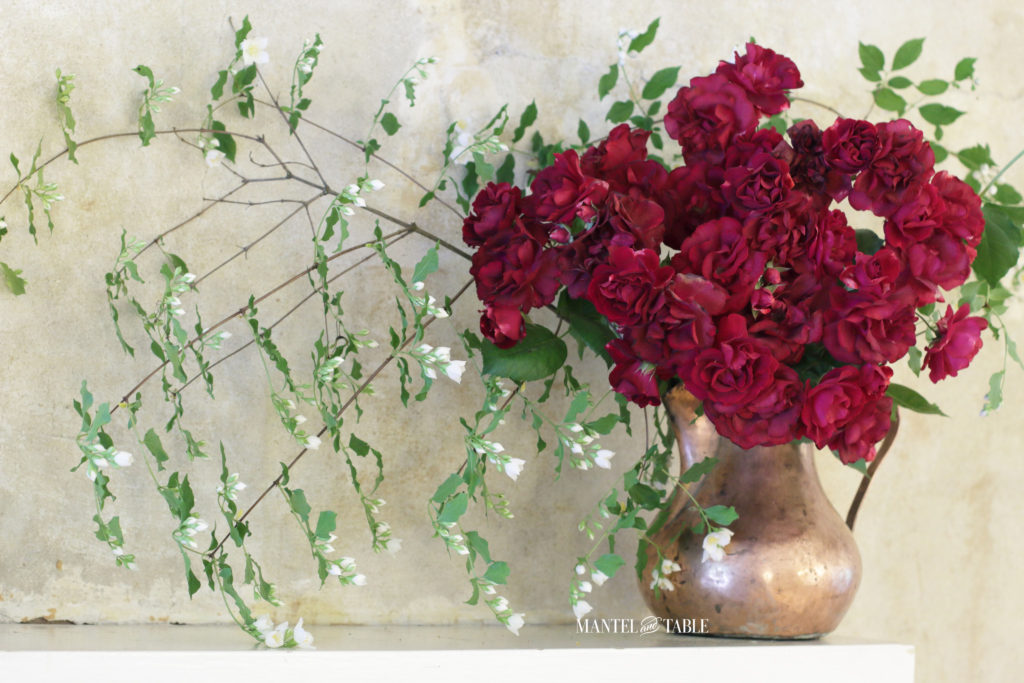 Red roses, mock orange branches in a copper pot on white mantel. Closeup